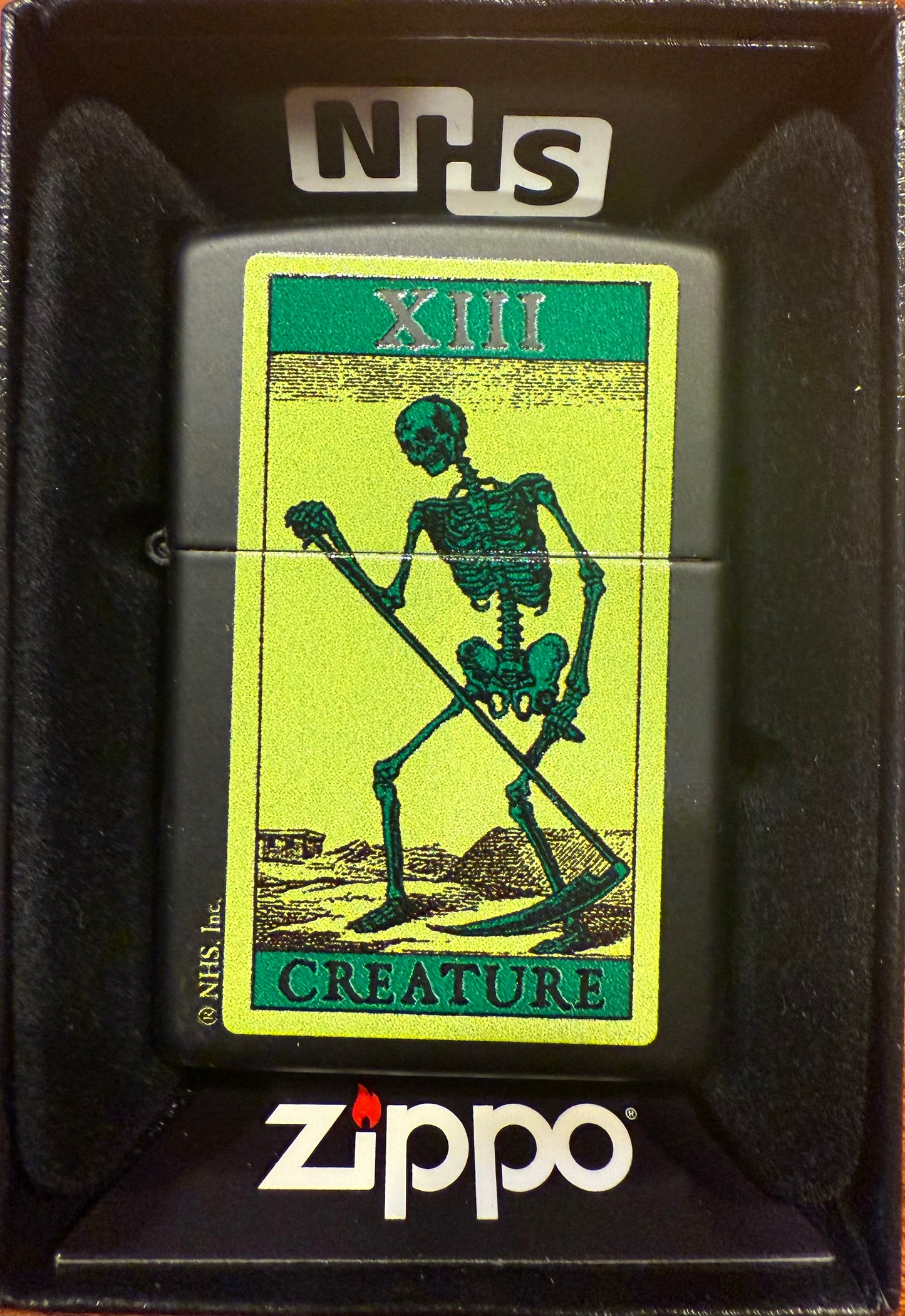 A Zippo lighter with a distinctive green Creature XIII Skeleton graphic on a black matte background, packaged in a gift box with the Zippo logo visible.