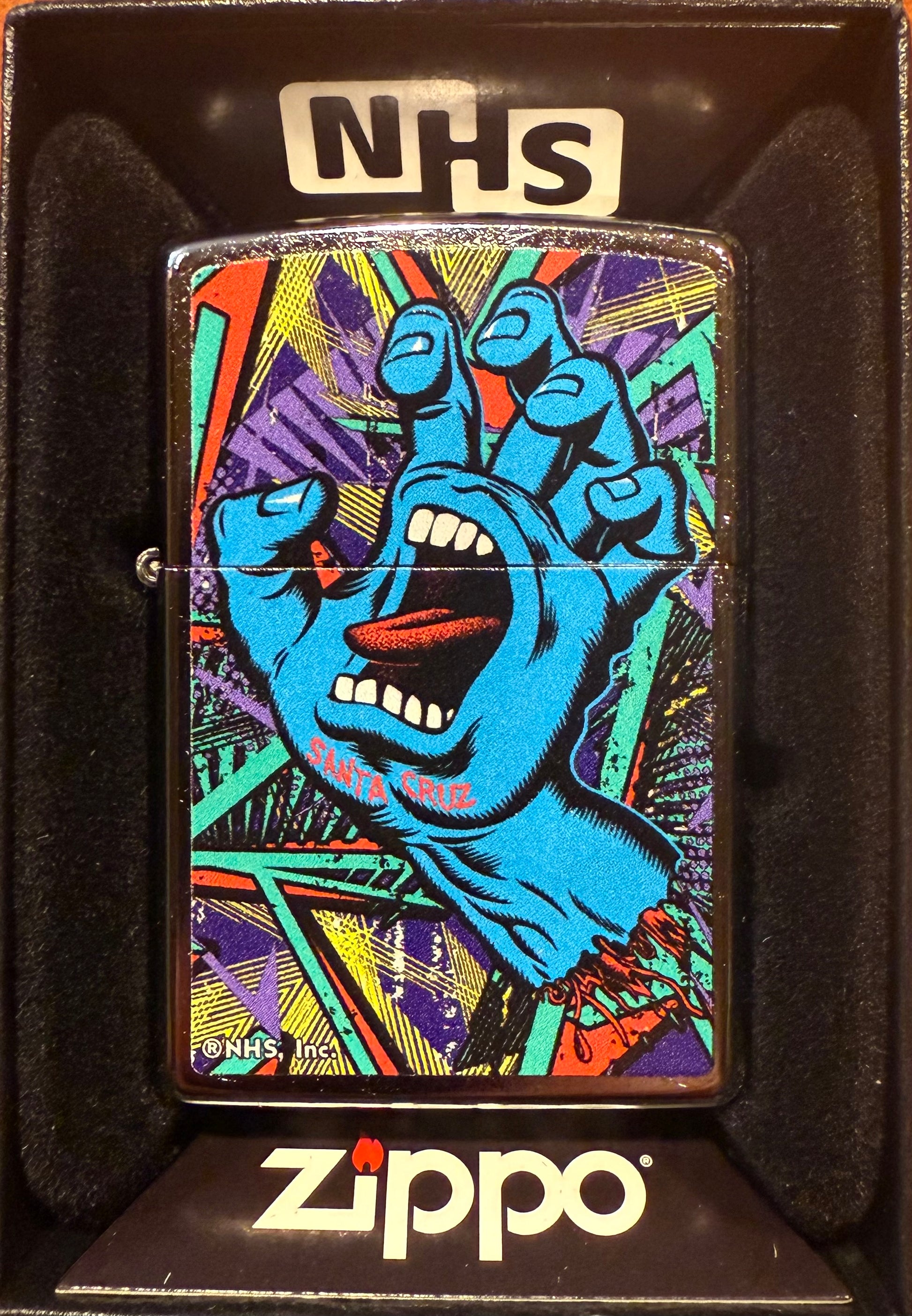 A Zippo lighter with a color(imaged) chrome finish featuring the iconic Screaming Hand graphic, inspired by Santa Cruz Skateboards' artwork, displayed in a black gift box with 'NHS' text visible.