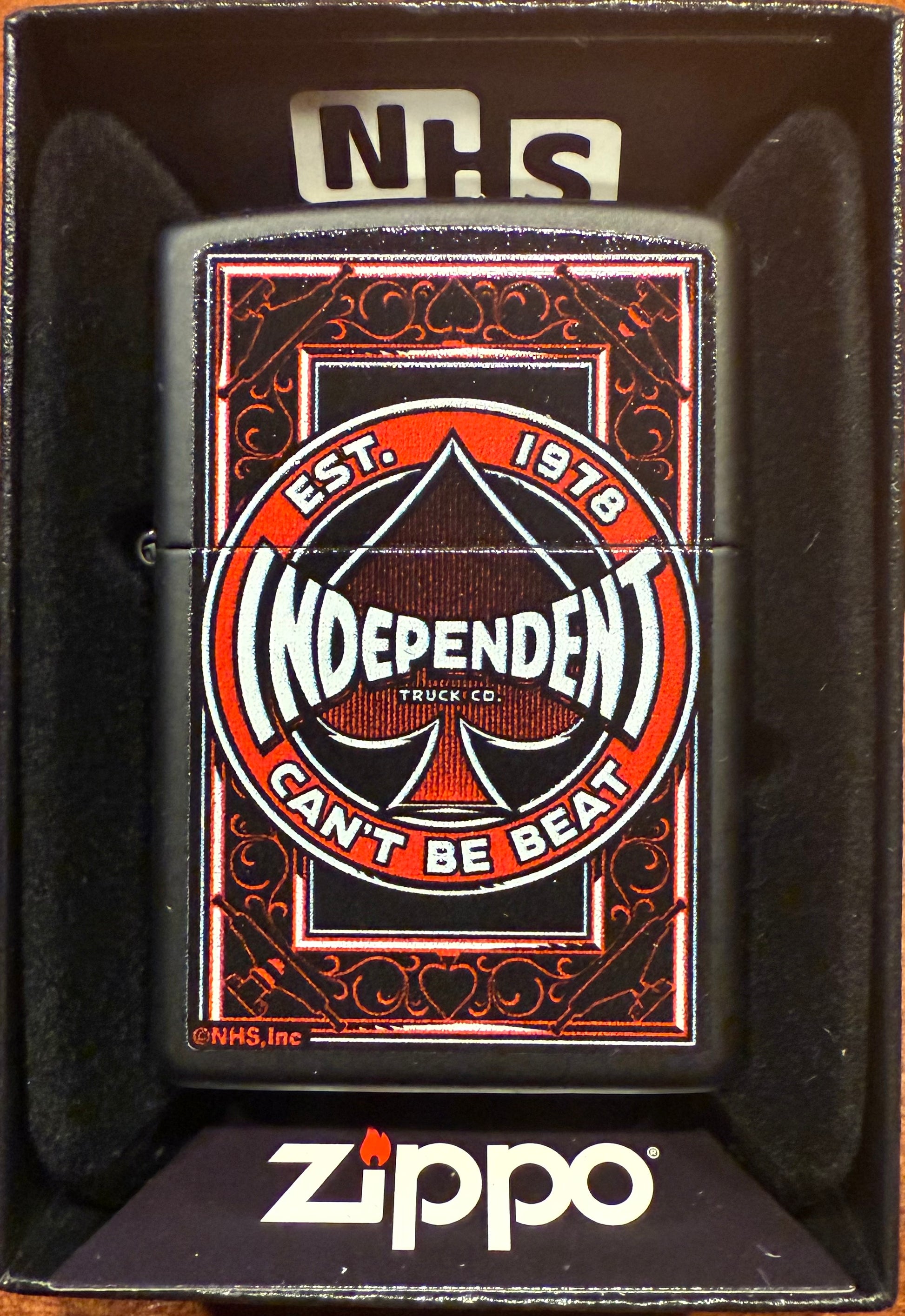 A Zippo lighter with a black matte finish featuring the Independent Spade logo in red and white, displayed in its packaging with the text 'NHS' on the top.