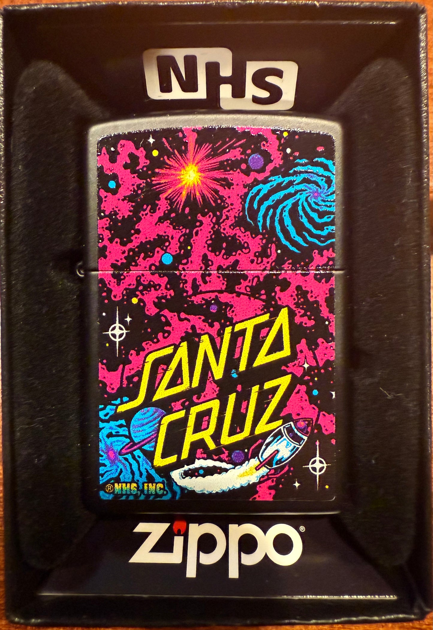 A Zippo lighter with a black matte finish featuring Santa Cruz Skateboards' iconic space-themed graphic, including stars and a logo.